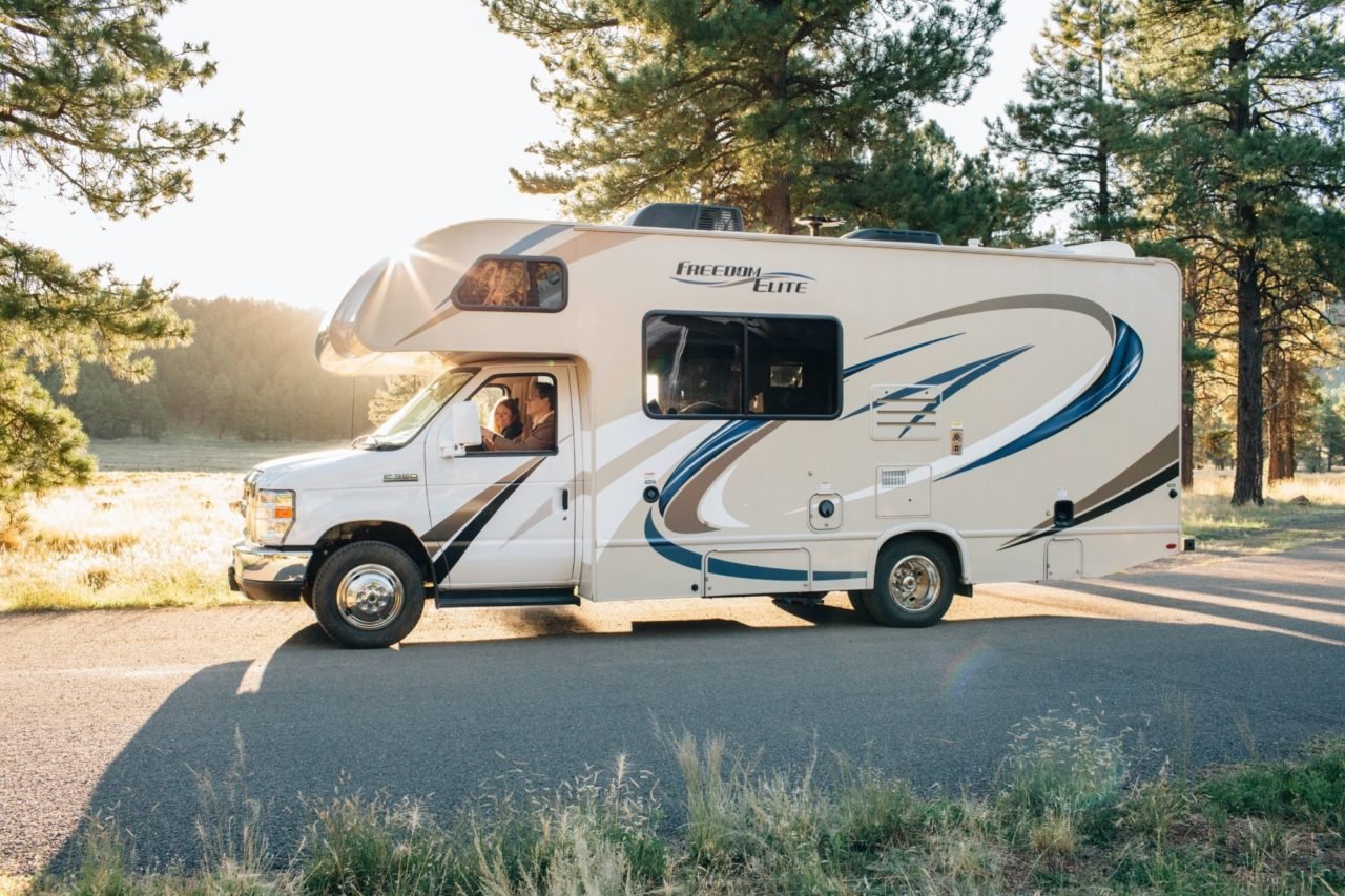 class c rv motorhome with two people inside and trees and sun in the background.
