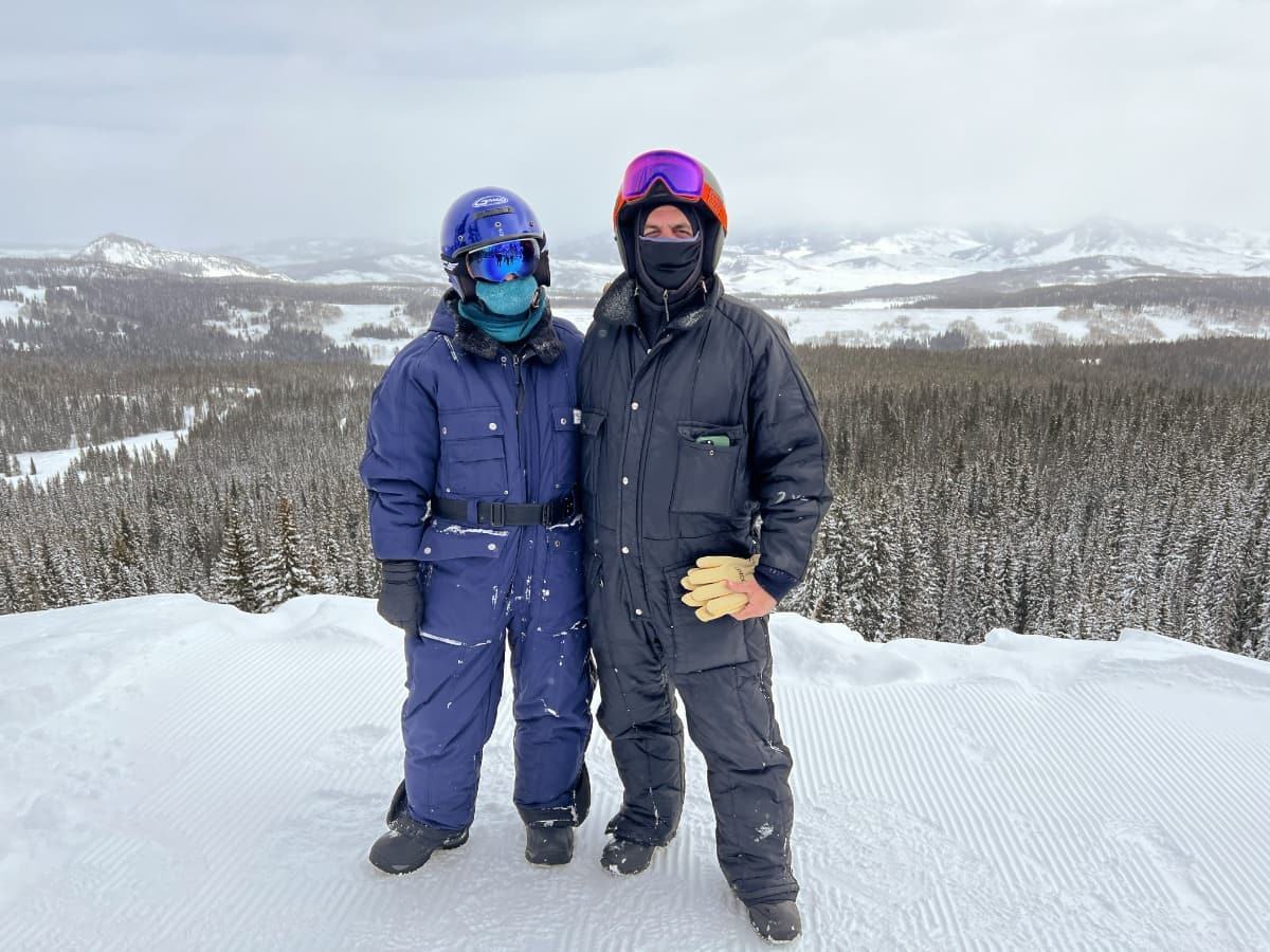 two people in snowsuits with snowy mountains in the background.