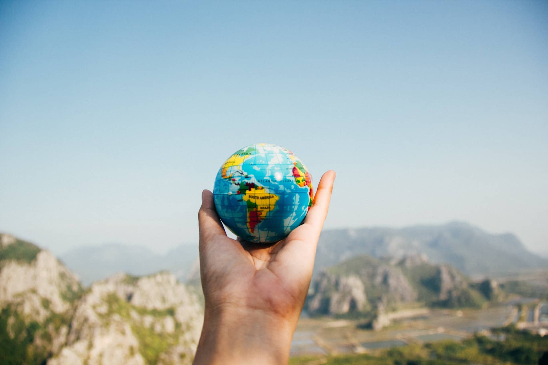 a hand holding a small globe with a mountain view in the background.